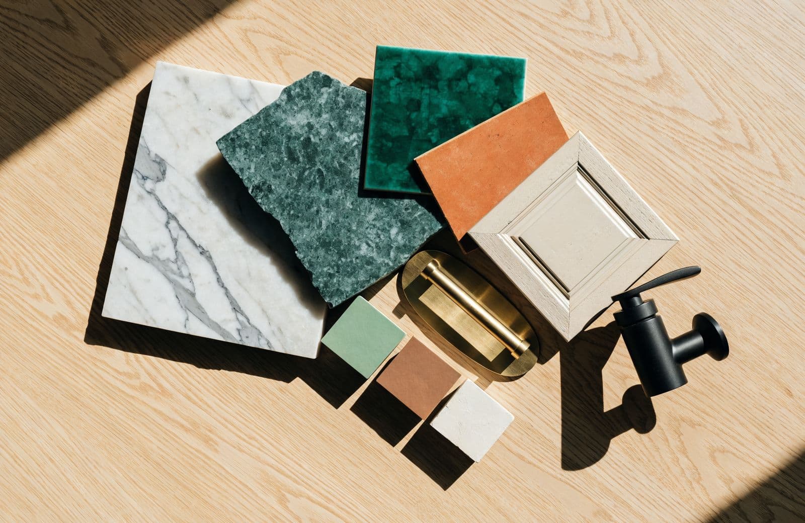 Material samples laid out on oak: marble, soapstone, emerald tile, terracotta tile, a cabinet door, a brass pull and three paint chips.